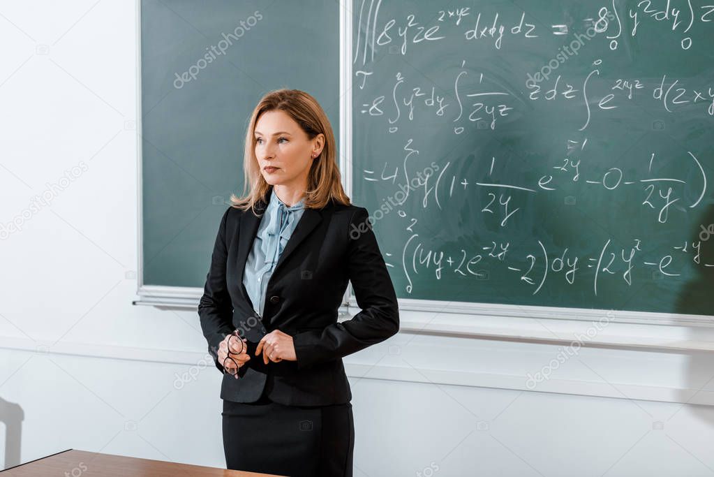 Female teacher standing in formalwear near chalkboard and holding glasses