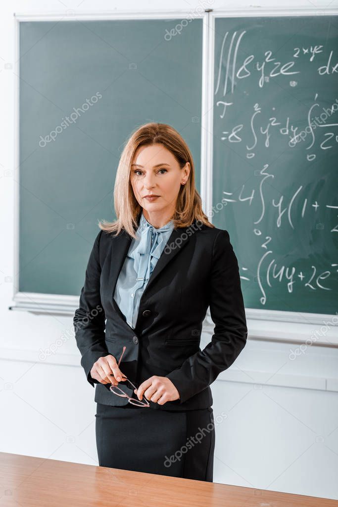 Beautiful teacher standing in classroom and holding glasses 