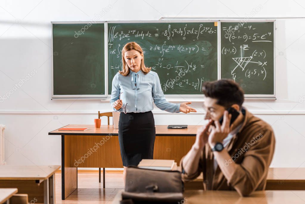 dissatisfied teacher looking at student who talking on smartphone in classroom 