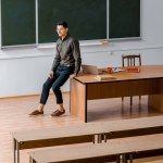 Männlicher Lehrer in formeller Kleidung, der wegschaut und auf dem Schreibtisch im Klassenzimmer sitzt