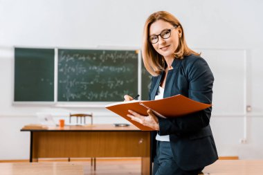 Not defterinde yazılı ve kamera sınıfta bakarak formal giyim güzel kadın öğretmen