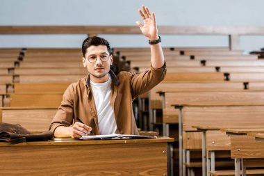focused male student in glasses sitting at desk and raising hand during lesson in classroom