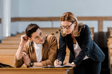 female teacher in glasses checking exam results of male student in classroom clipart