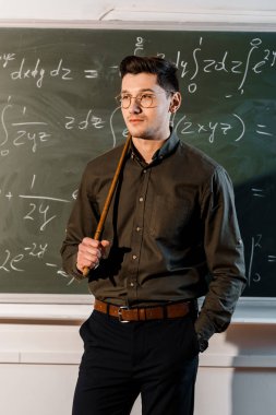 serious male teacher in formal wear holding wooden pointer in front of chalkboard with equations  clipart