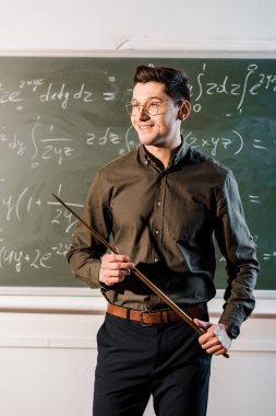 smiling male teacher in formal wear holding wooden pointer in front of chalkboard with equations  clipart