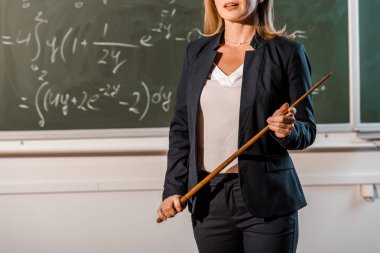 partial view of female teacher in formal wear with wooden pointer explaining mathematical equations in classroom clipart