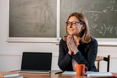 gülümseyen kadın öğretmen sınıfta boş ekran ile bilgisayar masa başında oturan