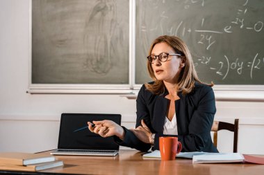 masada oturan ve sınıfta boş ekran ile laptop gösteren kadın öğretmen