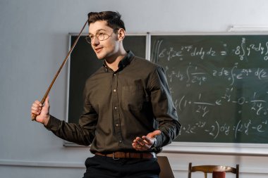 handsome male teacher in formal wear holding wooden pointer and explaining equations in class clipart