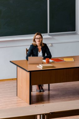 Masa başında oturan, kameraya bakıyor ve sınıfta dizüstü kullanarak kadın üniversite profesörü