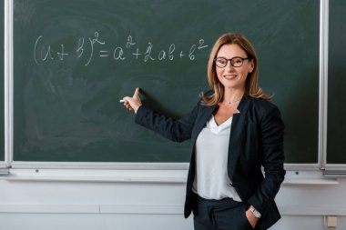 kadın öğretmen sınıfta kara tahta üzerinde matematiksel denklemi, parmakla işaret