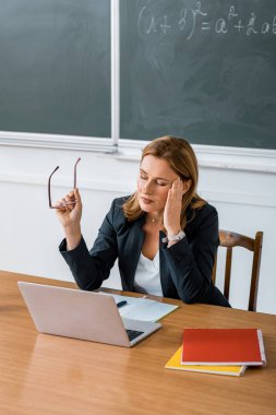 bilgisayar masasında oturan, gözlük tutan ve baş ağrısı sınıfta olan kadın öğretmen