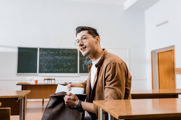 smiling male student in glasses sitting at desk and packing notebooks in classroom