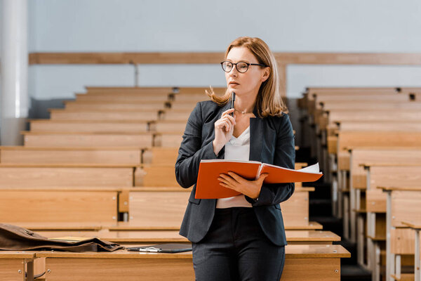 pensive female university professor holding journal and looking away in classroom