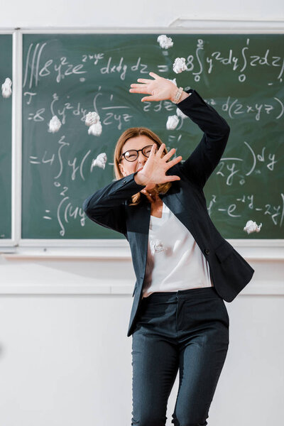 crumpled paper balls flying at frightened female teacher in classroom with chalkboard on background
