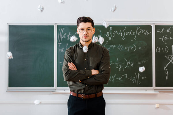 crumpled paper balls flying at male teacher with arms crossed in classroom with chalkboard on background