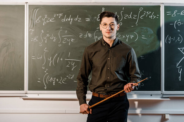 focused male teacher in formal wear looking at camera and holding wooden pointer in front of chalkboard with equations 