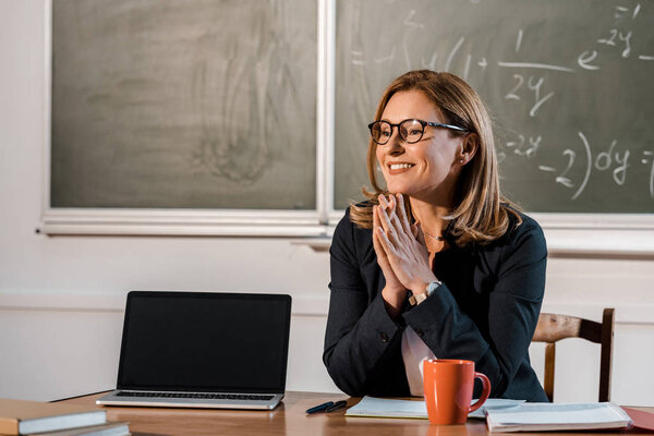 smiling female teacher sitting at computer desk with blank screen in classroom