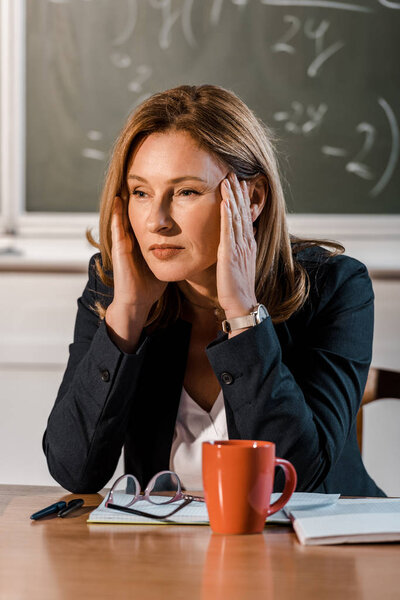 female teacher sitting at desk with hands on head and having headache in class