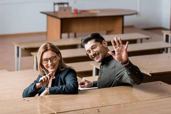 Mannelijke Student Zwaaien Camera Kijken Zit Klas Met Lachende Vrouwelijke — Gratis stockfoto