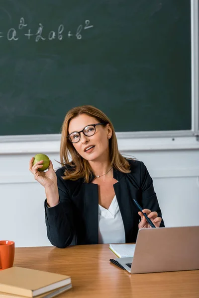 Guru Wanita Berkacamata Duduk Meja Komputer Dan Memegang Apel Kelas — Foto Stok Gratis