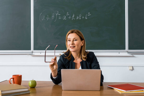 female teacher sitting at computer desk, holding glasses and talking in classroom 