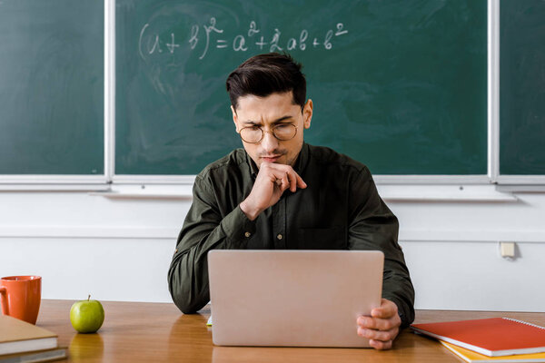 pensive male teacher in glasses sitting at desk and using computer in classroom