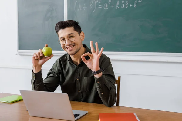 微笑的男老师在眼镜拿着苹果 并在教室的电脑桌上显示确定标志 — 图库照片