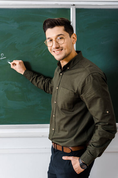 smiling male teacher looking at camera and writing on chalkboard in class