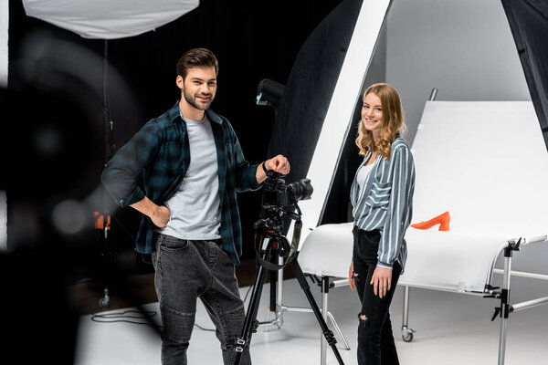 happy young photographers standing near professional photo equipment and smiling at camera in studio 