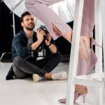 Tiro recortado de fotógrafo sorrindo sentado e fotografando modelo feminino elegante em estúdio