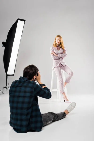 back view of photographer sitting and photographing beautiful female model posing in studio
