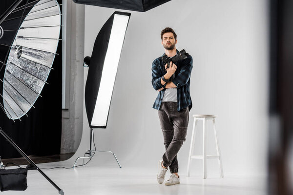 full length view of confident young photographer holding professional camera and looking at camera in photo studio 