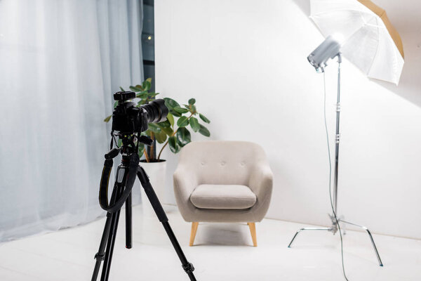 close-up view of professional photo camera on tripod, empty armchair and lighting equipment in studio