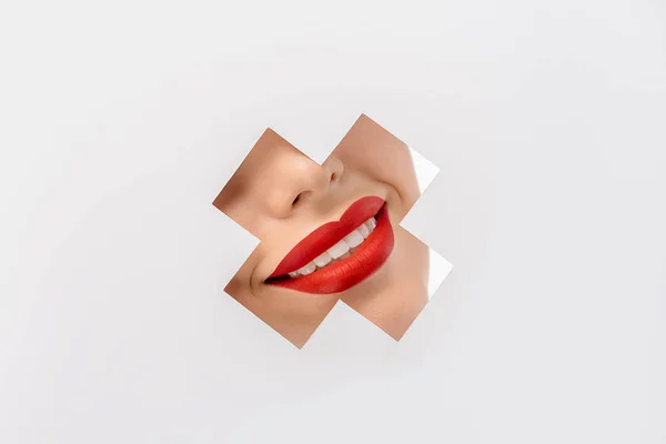 cropped shot of smiling young woman with red lips through cross shaped hole on white