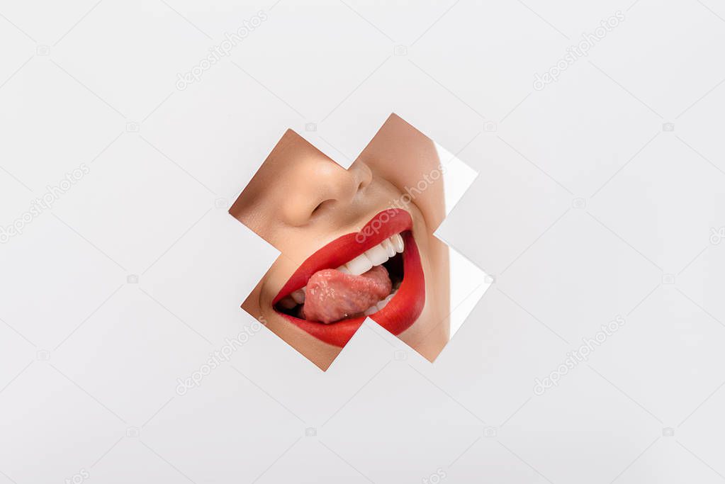 cropped shot of girl licking lips through cross shaped hole on white