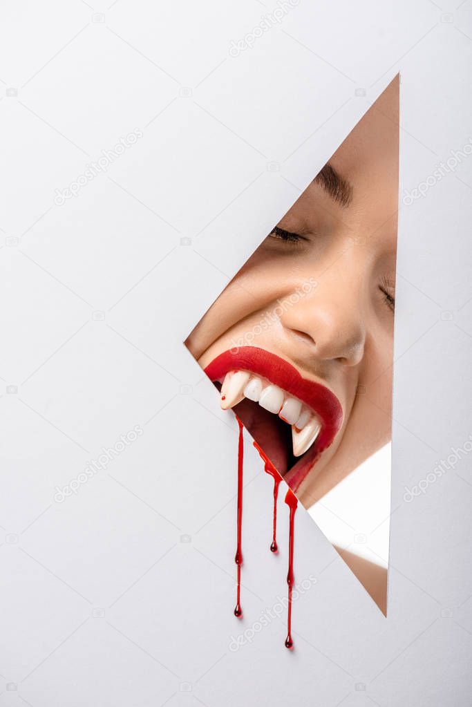 cropped shot of woman with vampire teeth and blood biting through hole on white 