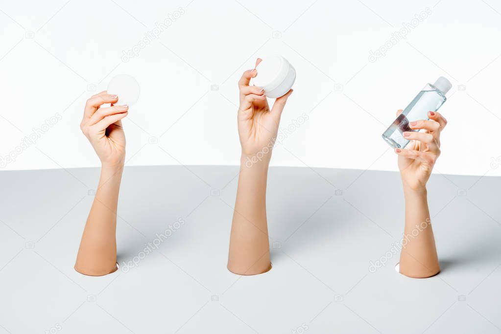 cropped shot of women holding cream, lotion and cotton pad through holes on white