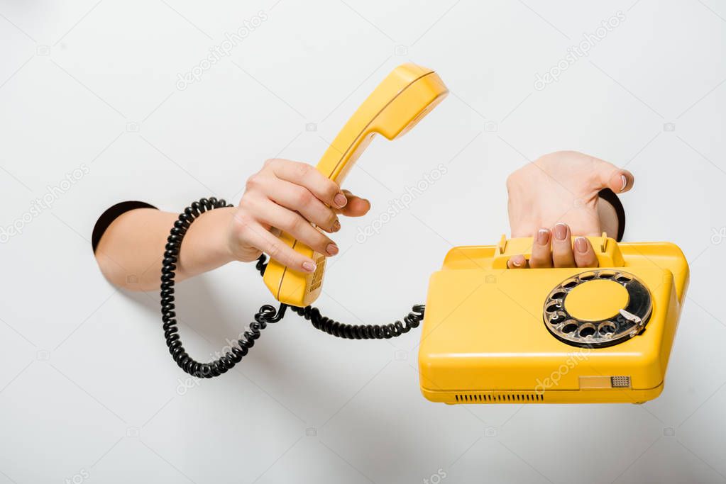 cropped image of woman holding retro yellow stationary telephone through holes on white