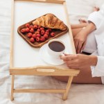 Imagem cortada de jovem segurando xícara de café enquanto sentado na cama com café da manhã na bandeja