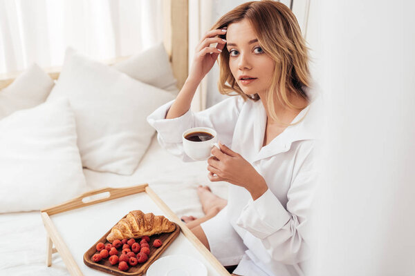 selective focus of young woman holding coffee cup while sitting on bed with breakfast on tray