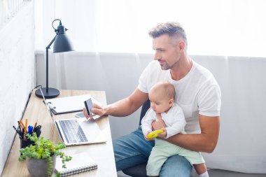 Kredi kartıyla online alışveriş ve bebek kızı evde tutan bilgisayar masa başında oturan yakışıklı baba