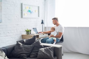 Masa başında oturan, dizüstü bilgisayar kullanarak ve bebek kızı modern apartman dairesinde tutan baba