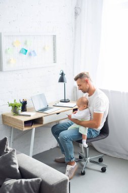 bilgisayar masasında oturan, smartphone kullanarak ve oturma odasında bebek kızı tutan baba