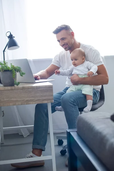 幸せな父持株赤ちゃん椅子に座っていると自宅のラップトップを使用して — ストック写真