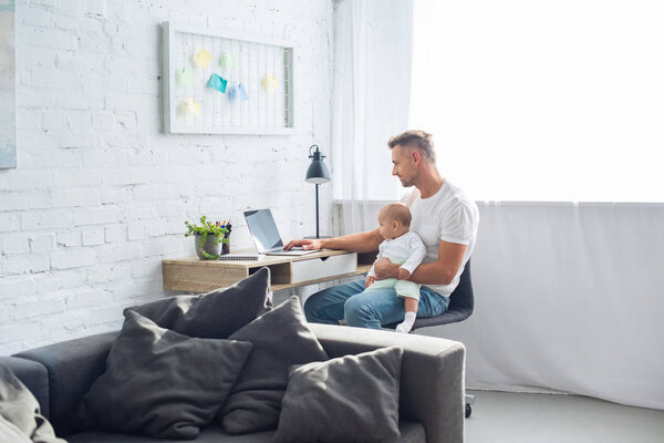 father sitting at desk, using laptop and holding baby daughter in modern apartment