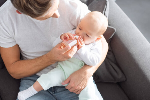 father sitting on couch and feeding baby daughter from bottle at home
