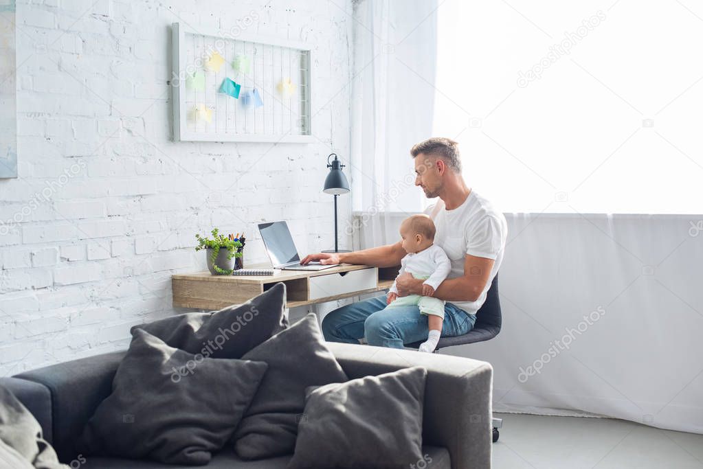 father sitting at desk, using laptop and holding baby daughter in modern apartment