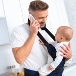 Happy father holding infant daughter in baby carrier and talking on smartphone