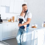 Serious father holding infant daughter in baby carrier and talking on smartphone